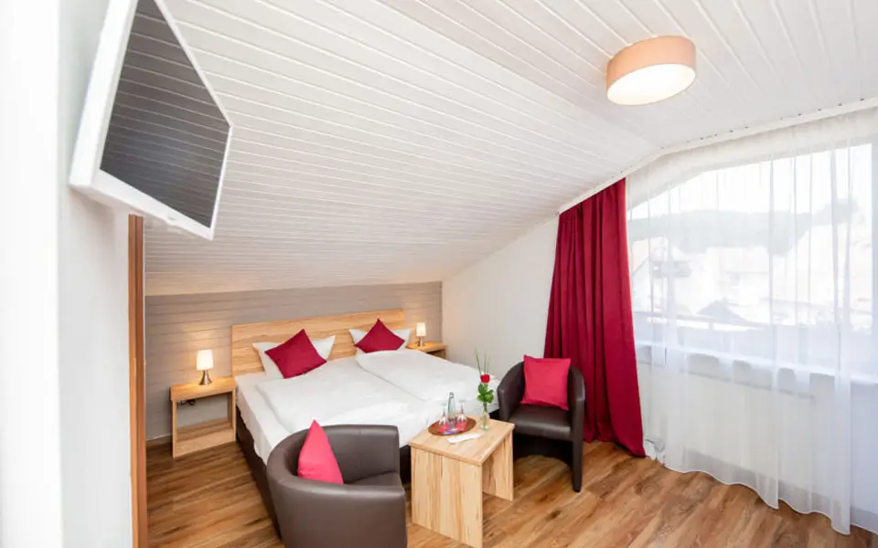 Dreibettzimmer mit zwei Einzelbetten und einem Einzelbett, zwei Sesseln mit roten Kissen, Holztisch, Nachttischen mit Lampen und großem Fenster mit rotem Vorhang