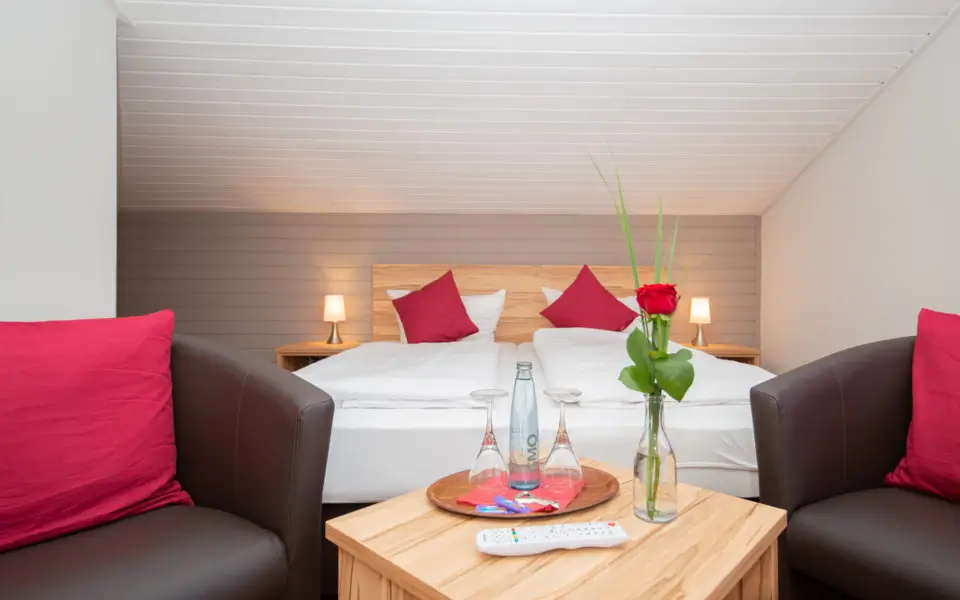 Hotelzimmer mit Doppelbett, zwei Sesseln mit roten Kissen, einem Holztisch mit Wasserkaraffe, Gläsern, Fernbedienung und einer roten Rose in einer Glasvase
