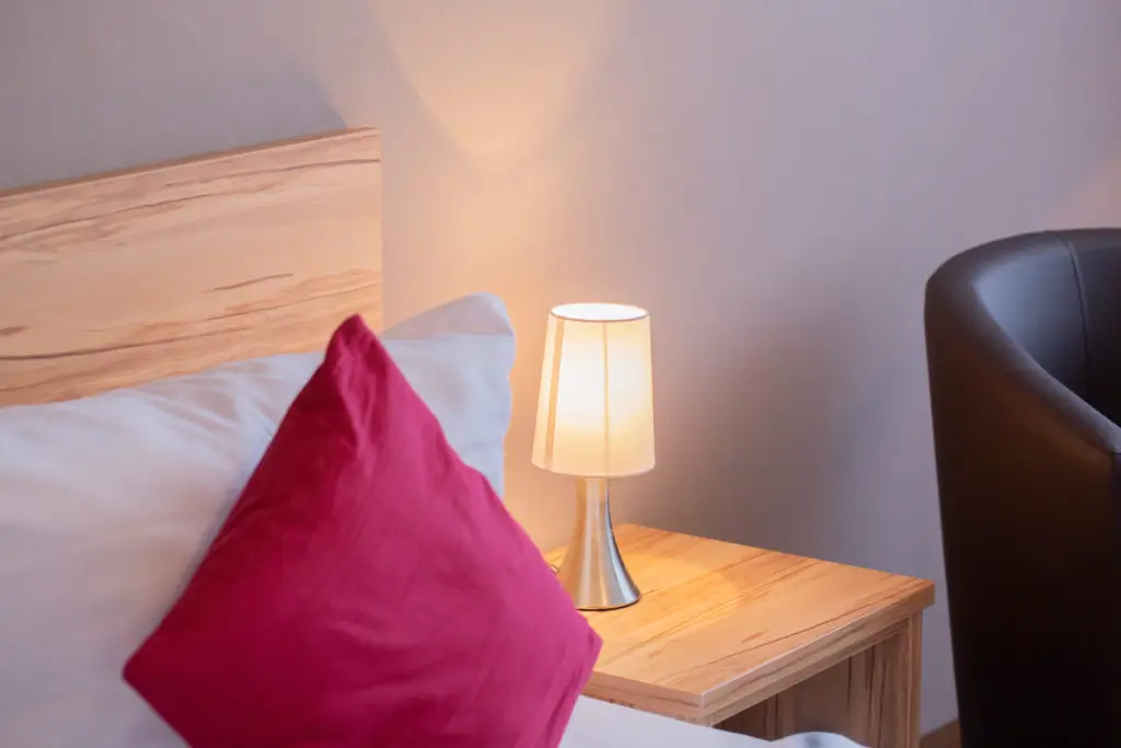 Bett mit weißem Kissen und rotem Zierkissen neben einem Holznachttisch mit einer kleinen Lampe, rechts ein schwarzer Sessel
