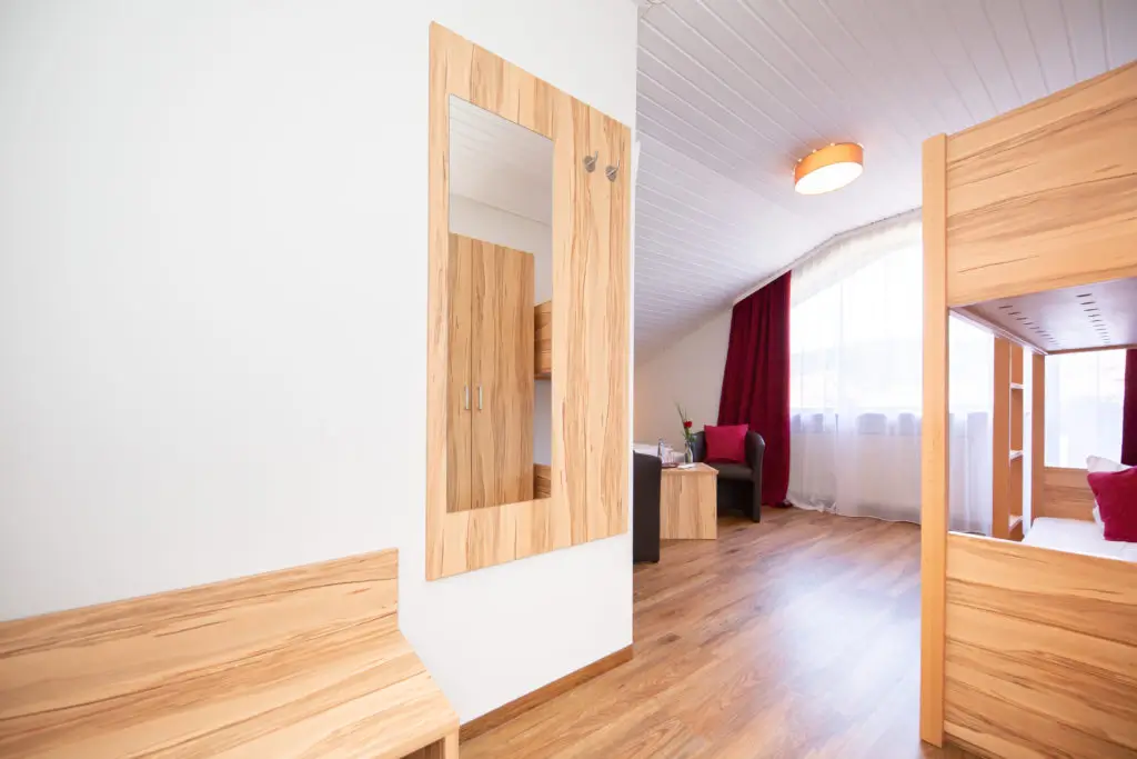 Flur mit Holzbank, großem Wandspiegel und Blick in ein helles Zimmer mit Fenster, rotem Vorhang und Sitzmöbeln und Etagenbett