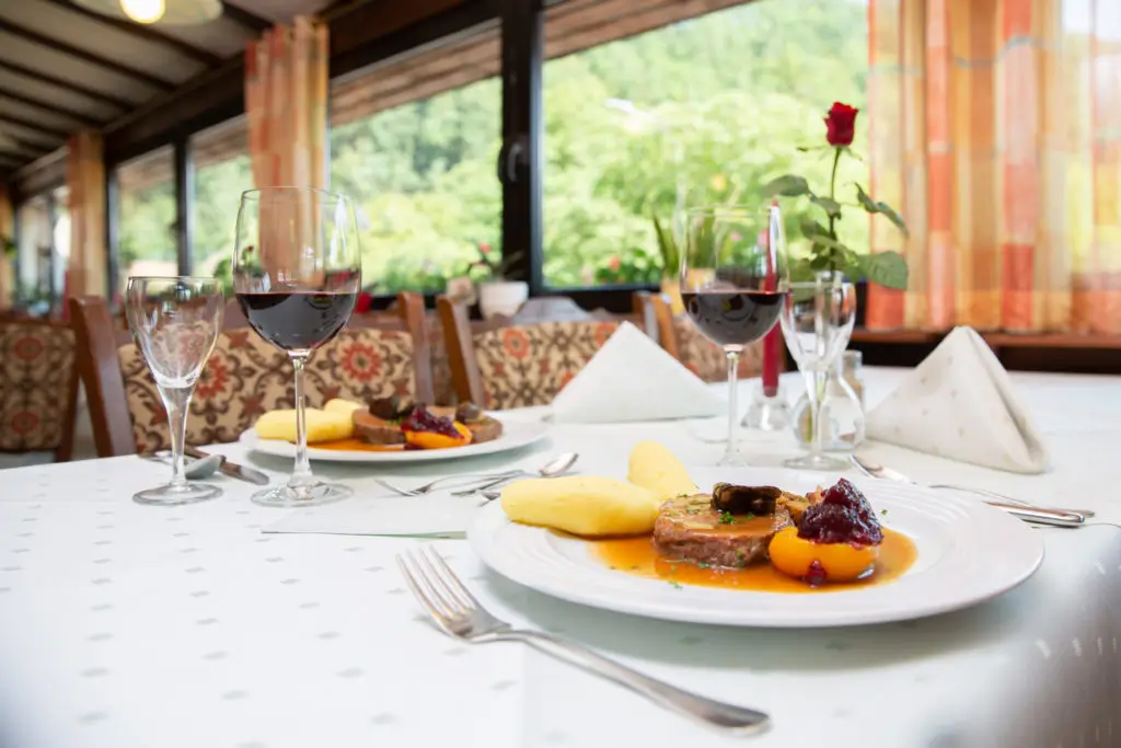 Tisch im Restaurant mit gedecktem Teller, Rotweinglas, Wasserglas und Servietten vor großen Fenstern