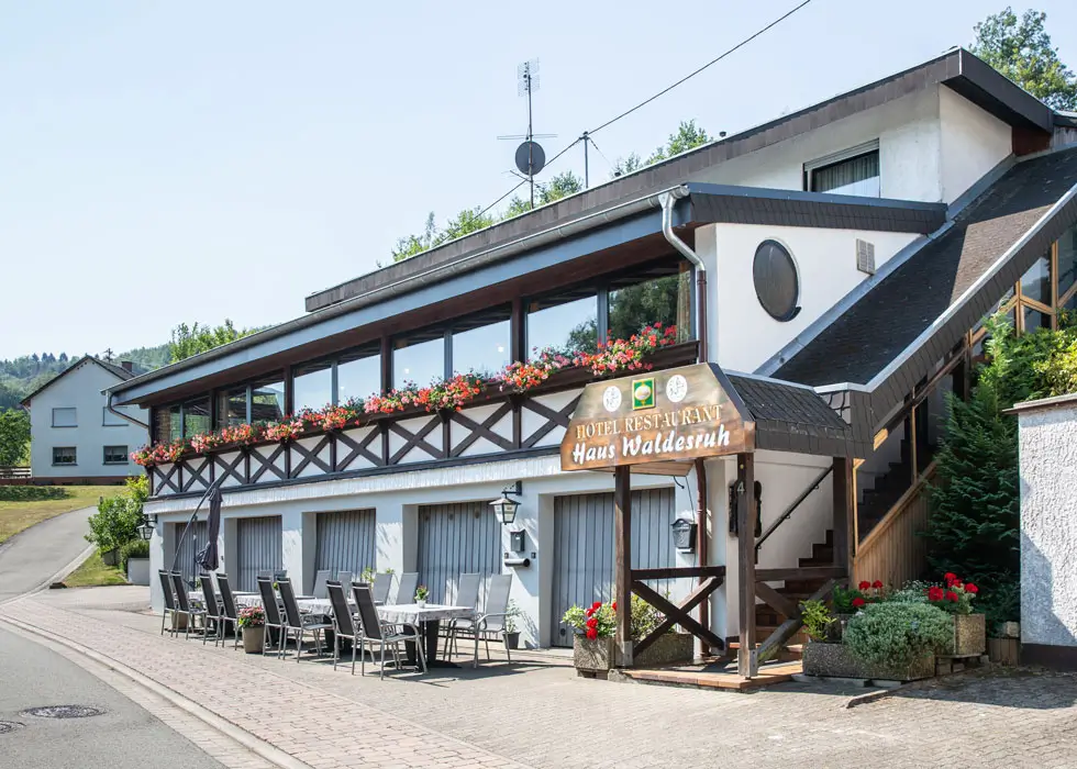 Hotel-Restaurant Waldesruh mit Terrasse, Tischen und Stühlen, Holzgeländer mit Blumenkästen und Außentreppe, umgeben von grüner Landschaft