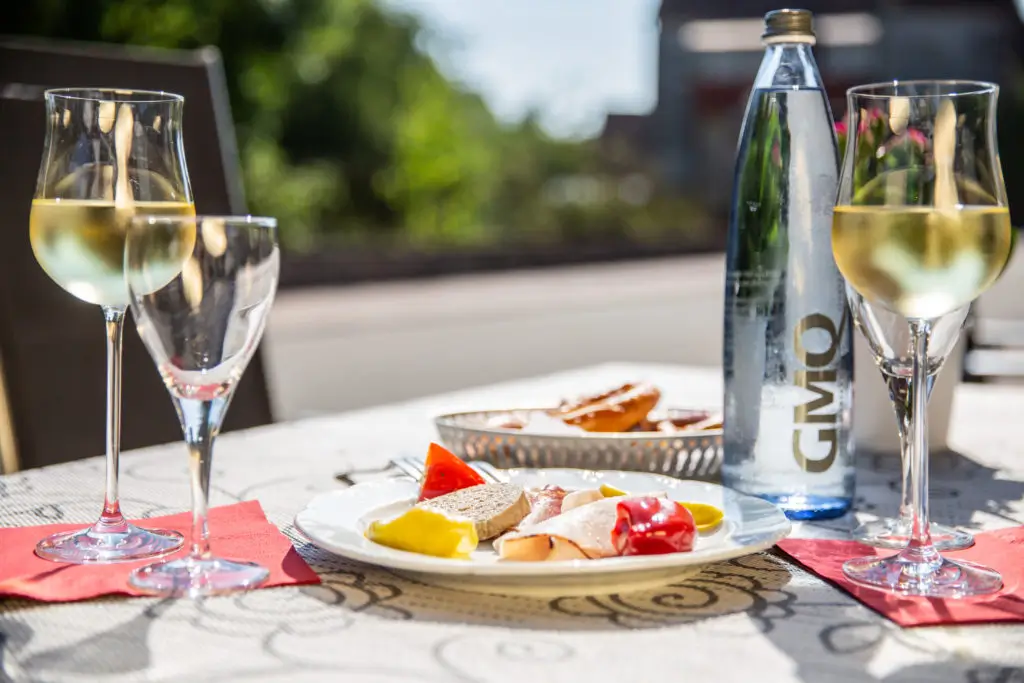 Tisch mit zwei Gläsern Weißwein, einer Flasche Mineralwasser und einem Teller mit verschiedenen kleinen Speisen, darunter Käse und Paprika