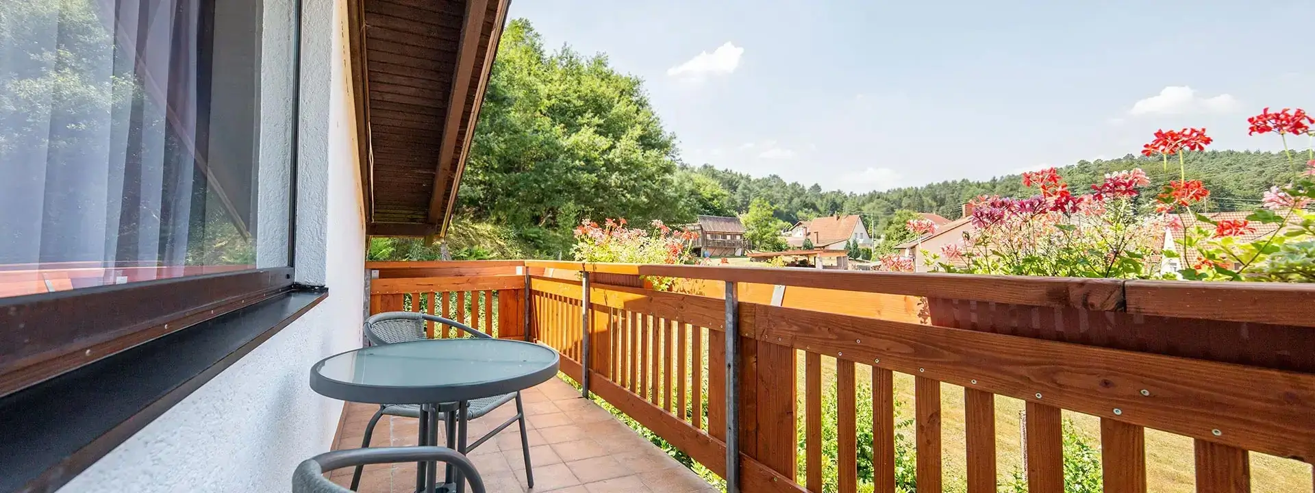 Balkon mit zwei Stühlen, kleinem runden Tisch und Holzgeländer mit Blick auf grüne Bäume und Häuser im Hintergrund