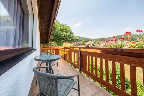 Balkon mit zwei Stühlen, kleinem runden Tisch und Holzgeländer mit Blick auf grüne Bäume und Häuser im Hintergrund