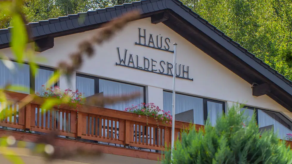 Blick auf das mehrstöckige Haus Waldesruh mit Balkon, umgeben von Bäumen und Sträuchern
