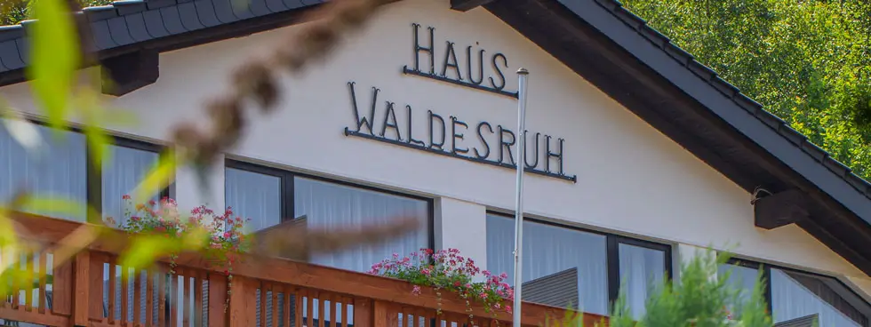 Blick auf das mehrstöckige Haus Waldesruh mit Balkon, umgeben von Bäumen und Sträuchern