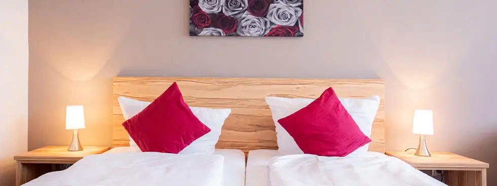 Doppelbett mit 2 Matratzen, je einer weißen Bettdecke, je ein rotes Kissen, Holzkopfteil, zwei Nachttische mit Lampen und ein Bild mit Rosen an der Wand