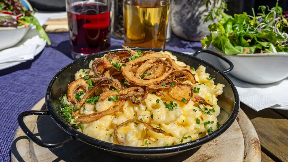 Schwarze Pfanne mit Käsespätzle, garniert mit Röstzwiebeln und Schnittlauch, auf Holzunterlage, im Hintergrund Salat und Getränke.