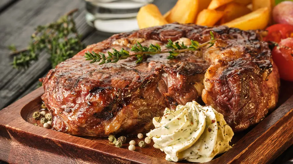 Gegrilltes Rumsteak auf Holzbrett mit Kräuterbutter, Pommes und Tomatenscheiben im Hintergrund.