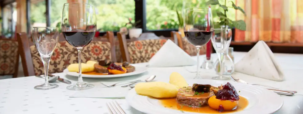 Tisch im Restaurant mit gedecktem Teller, Rotweinglas, Wasserglas und Servietten vor großen Fenstern