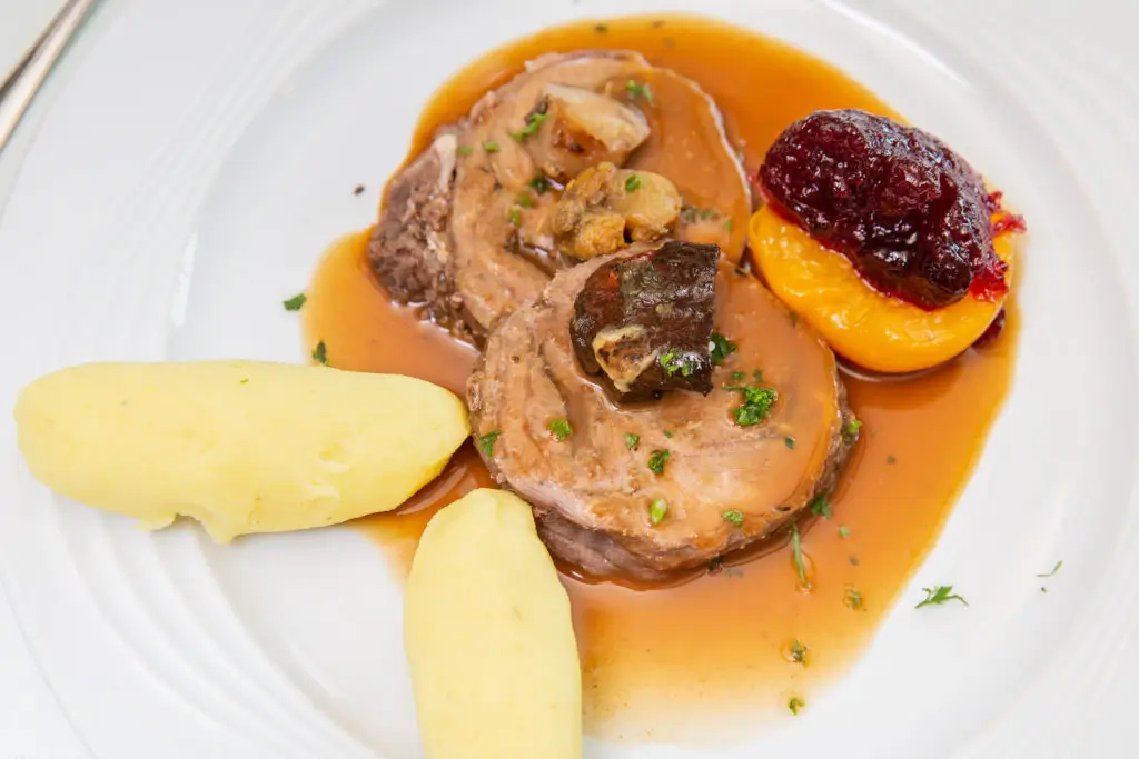 Zwei Schweinemedaillons mit brauner Sauce, dazu zwei Nocken Kartoffelpüree und eine halbe Pfirsichhälfte mit roter Sauce auf weißem Teller.