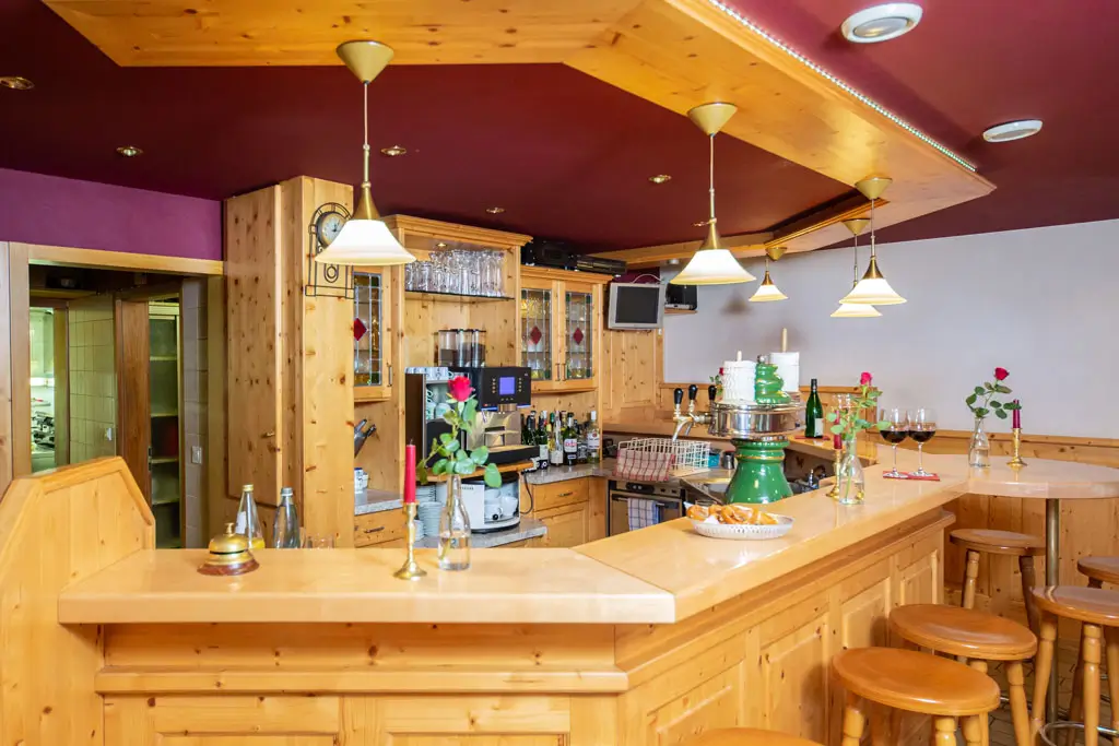 Holztheke mit Barhockern, mehreren Weingläsern und einzelnen roten Rosen in kleinen Vasen in einem Restaurant mit Holzdecke und roten Wänden.