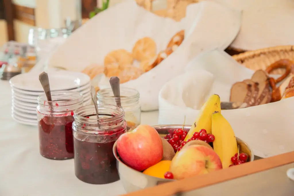 Frühstückstisch mit zwei Marmeladengläsern, einem Obstkorb mit Äpfeln, Banane und roten Johannisbeeren sowie einem Brotkorb im Hintergrund.