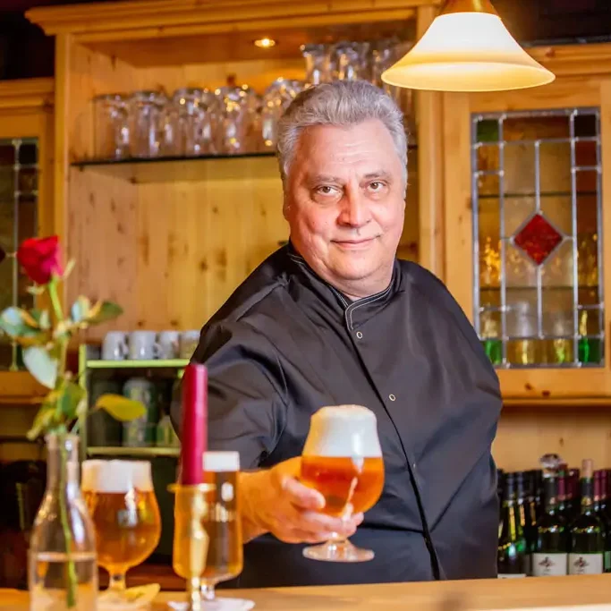 Kellner in schwarzem Hemd reicht ein Glas Bier an der Holztheke eines Gasthauses mit Glasregal und dekorativen Fenstern im Hintergrund.