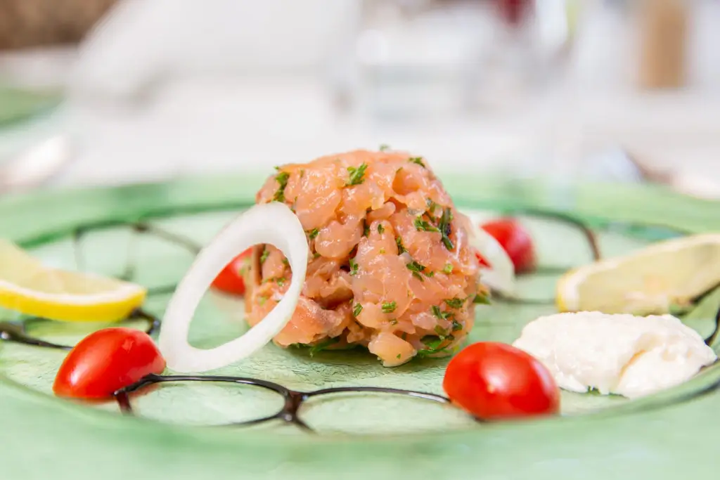 Auf einem grünen Teller liegt eine Kugel aus gehacktem Lachs, umgeben von Zwiebelringen, Kirschtomaten und einem Klecks Frischkäse.