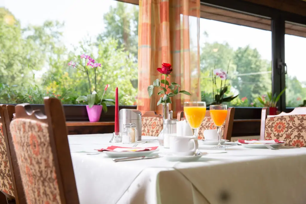 Eingedeckter Tisch mit weißen Tischdecke, zwei Gläsern Orangensaft, einer roten Rose in Vase und Stühlen mit gemustertem Polster vor großen Fenstern mit Blick ins Grüne.