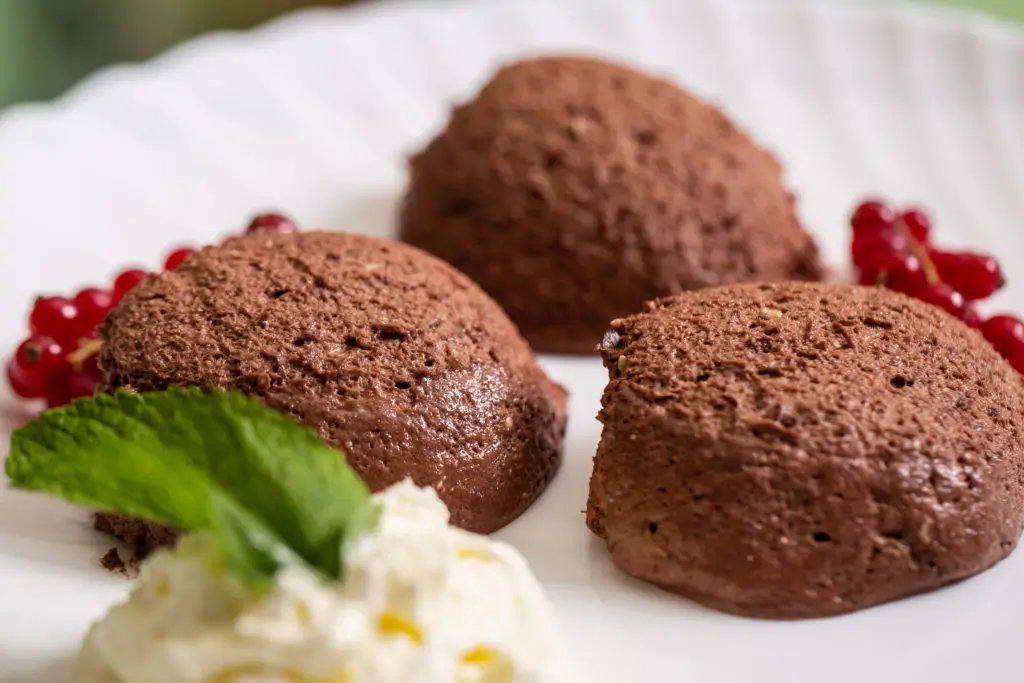 Drei Schokomousse-Portionen auf weißem Teller, garniert mit roten Johannisbeeren und einem Klecks Sahne mit Minzblatt.
