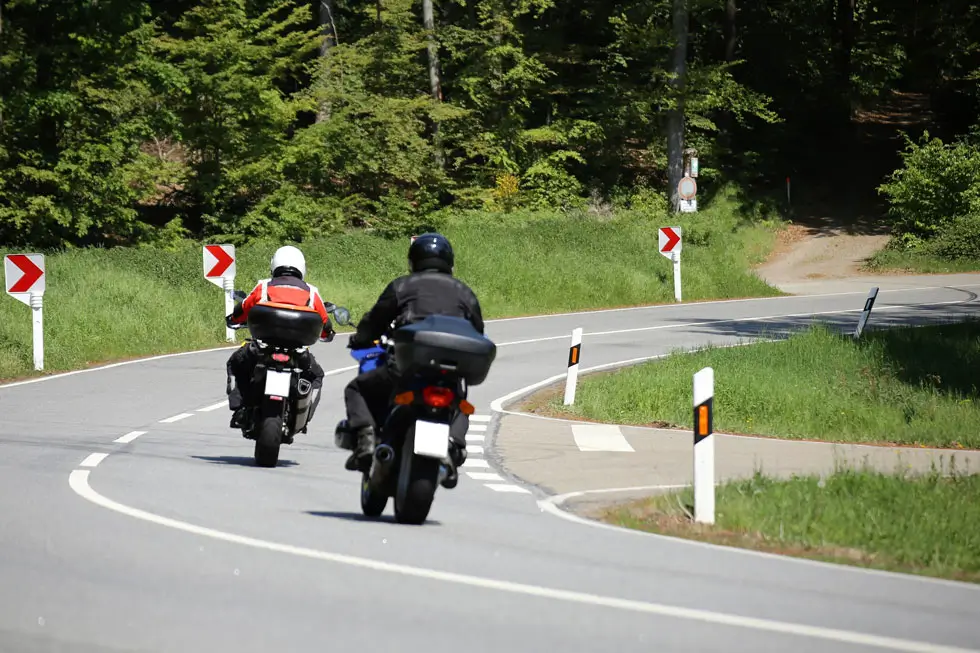 Zwei Motorradfahrer mit Helmen fahren auf einer kurvigen Landstraße mit Leitpfosten und Wald im Hintergrund